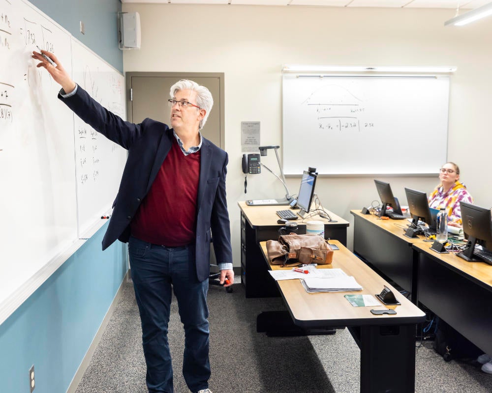 A professor stands at the front of a classroom, teaching data analytics equations to a room of students.