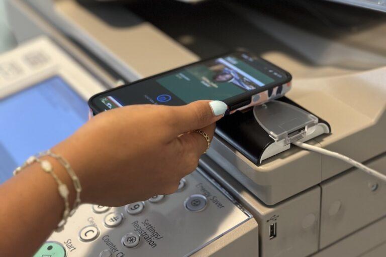 A student holds their phone up to an RFID reader on a copy machine. The phone screen shows the student's mobile ID card.